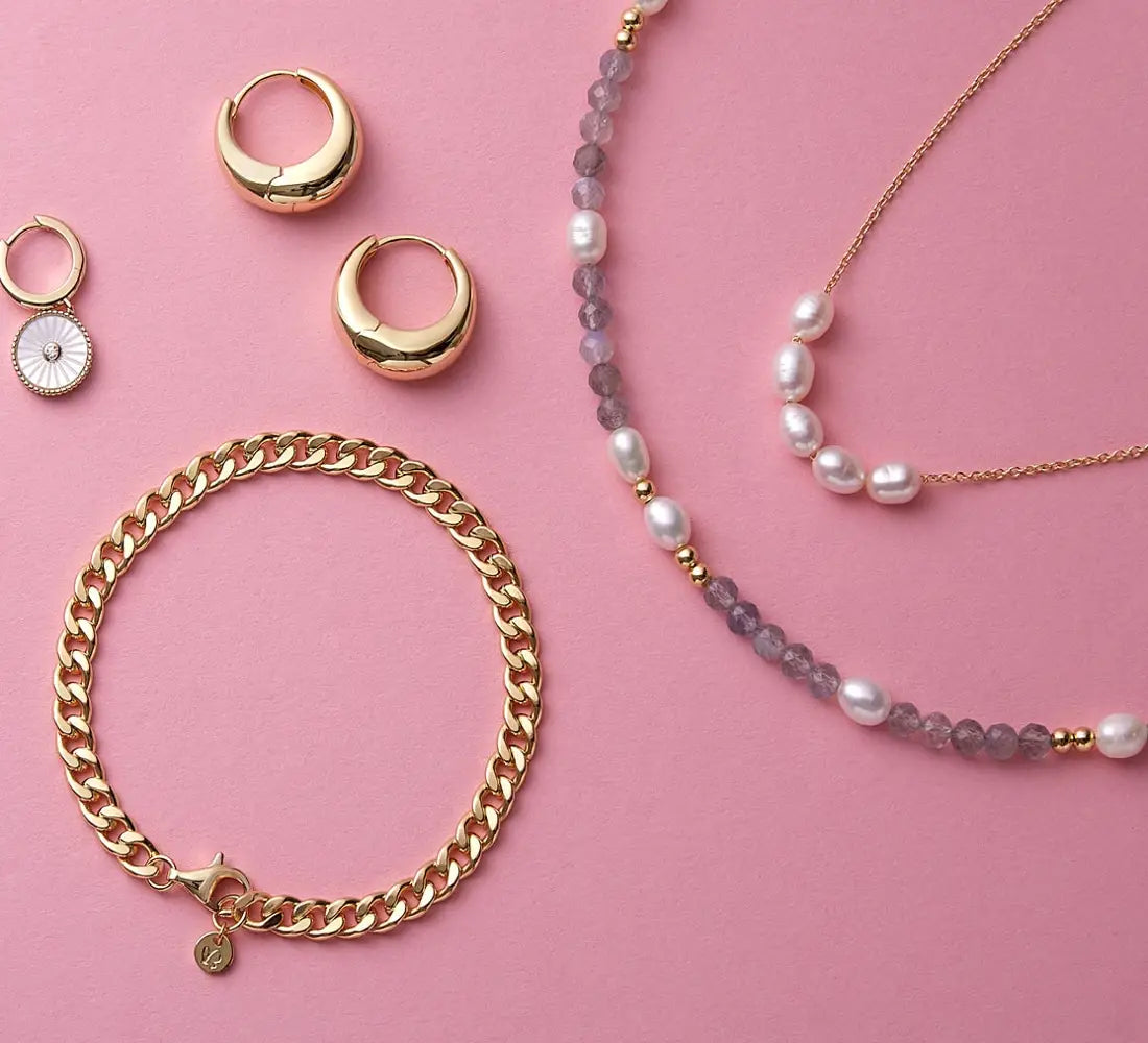 Goldfarbene Ohrringe, Armband und Perlenkette auf rosafarbenem Hintergrund